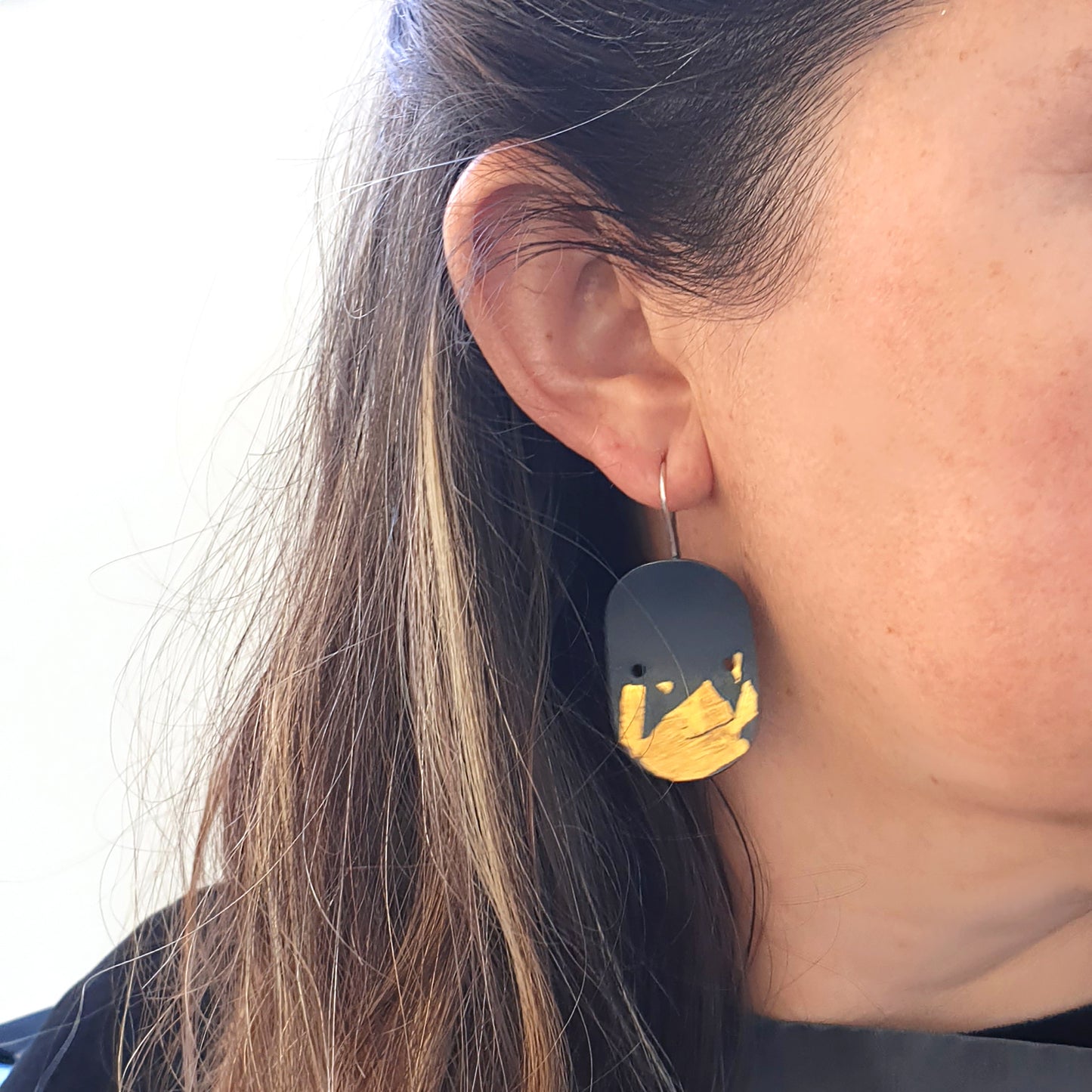 The Curva.dos Earrings by FR | Art Jewellery feature an oval shape with a black base and gold foil accents, highlighting the Keum Boo technique. Each earring comes with a sterling silver hook for fastening and a minimalist design that includes two small white dots near the top.